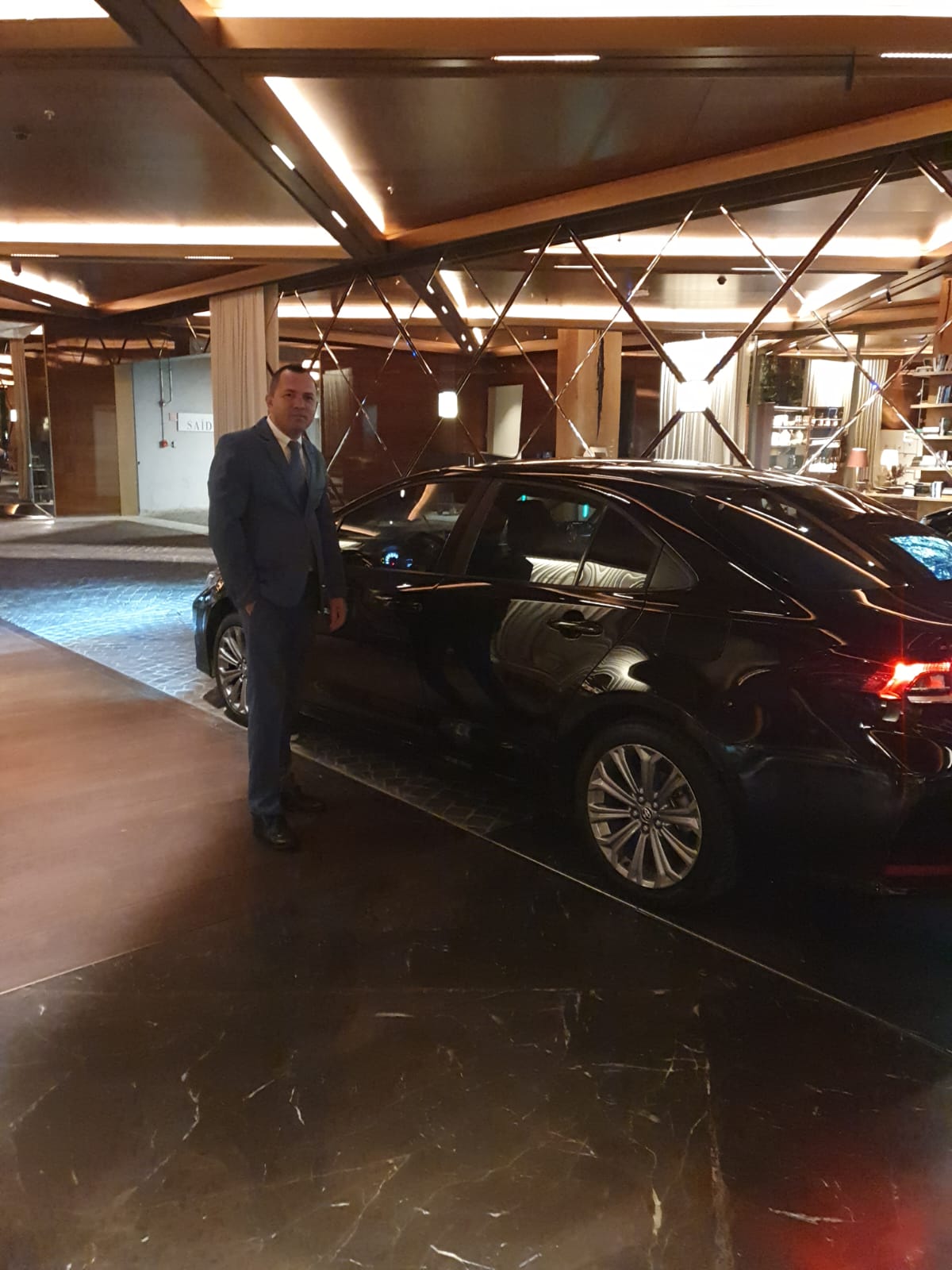 Motorista executivo de terno ao lado de sedã premium em lobby de hotel